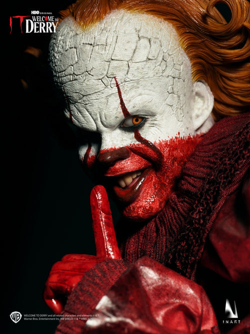 A close-up promotional shot of a Pennywise 'Welcome to Derry' collectible figure or statue from the movie IT, showing a detailed painted face with white makeup, red paint streaks, orange hair, and a red ruffled collar against a dark background.