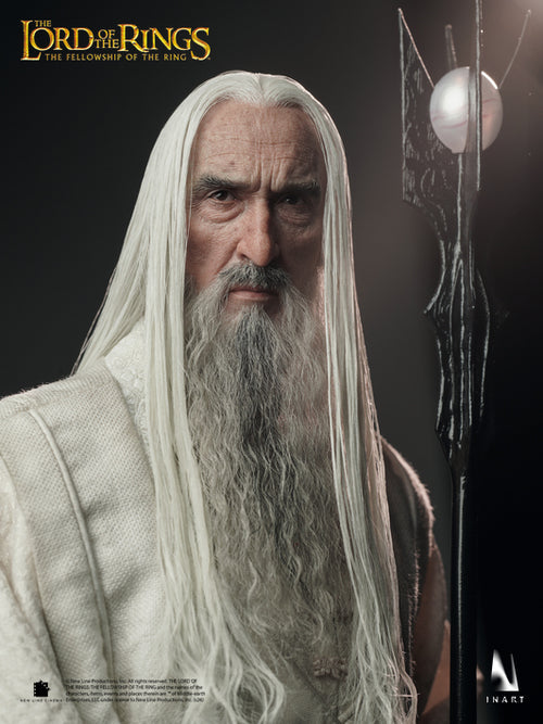 A high-end collectible figure of Saruman from The Lord of the Rings, featuring long white hair and beard, wearing a textured white robe, posed next to his staff against a dark studio background.