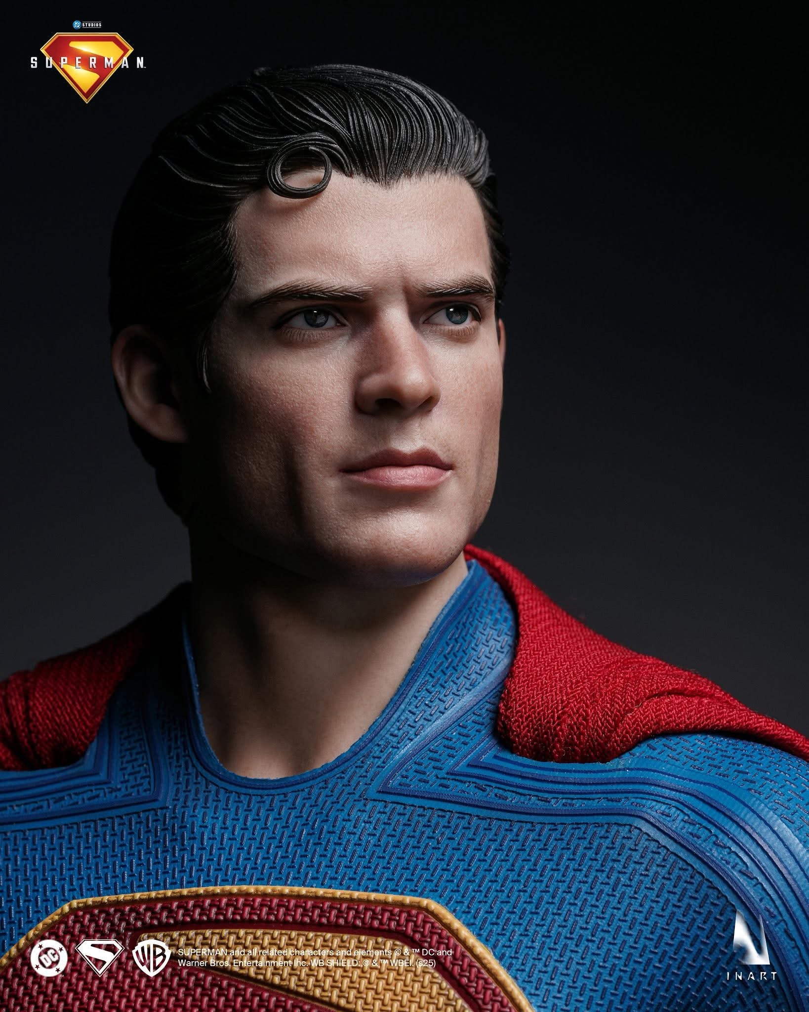 Superman collectible figure bust with detailed sculpted face, dark hair, textured blue suit, and red cape against a dark studio background. Superman logo and small brand marks are visible on the image.