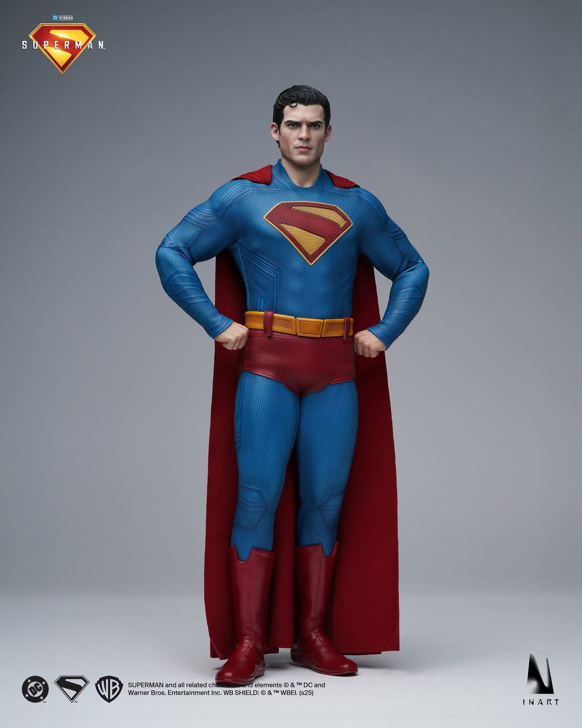 A Superman collectible figure posed standing with hands on hips, wearing a blue suit with a red cape and red-and-yellow chest emblem, photographed on a neutral gray studio background. Superman and DC Entertainment logos are visible in the top left, and small copyright marks appear along the bottom.